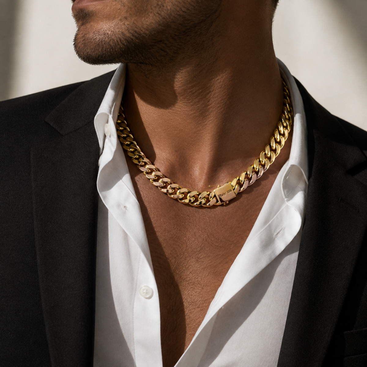 A close-up of a man wearing a thick gold Cuban link chain necklace over an open white dress shirt and black blazer.