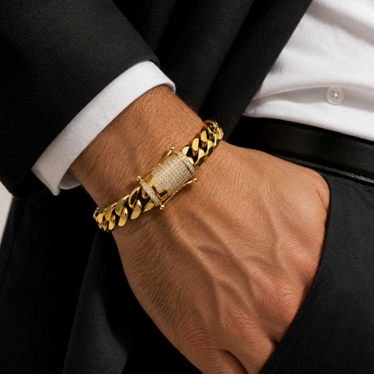 A close-up of a man's hand wearing a chunky gold Cuban link chain bracelet with a central diamond-encrusted clasp, styled with a black suit jacket and white dress shirt.